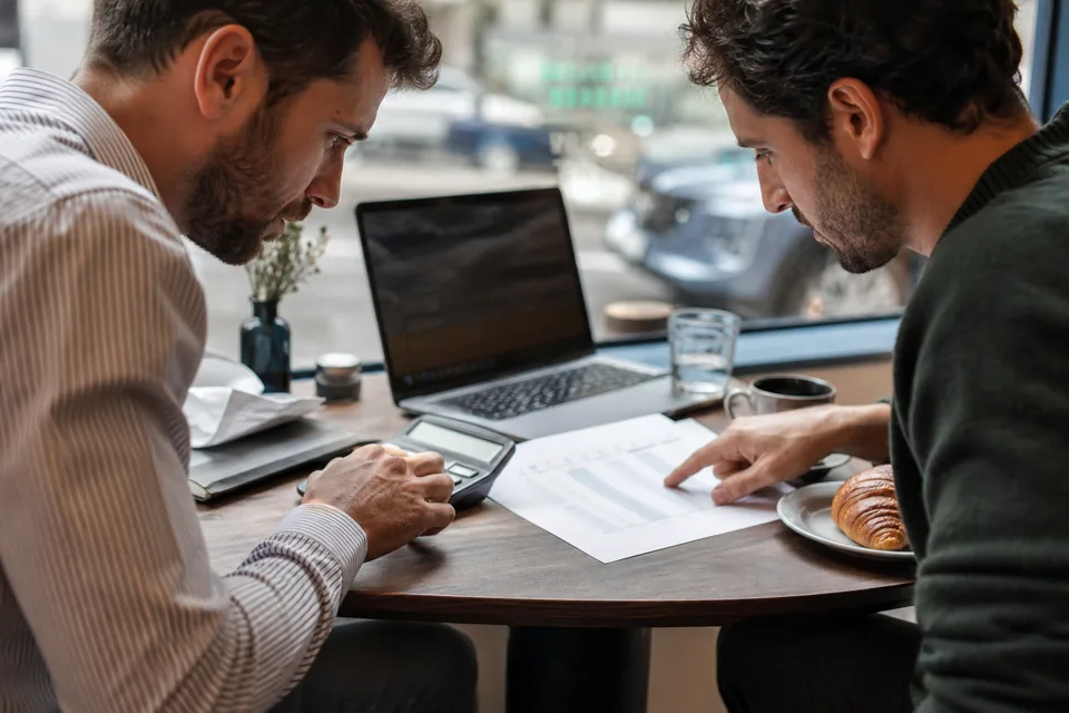 Two founders at a cafe table comparing a printed monthly software budget with a calculator
